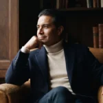A composed Latino man in his early fifties in a navy blazer and cream roll-neck sits in a leather chair in a wood-panelled study, looking toward the window while designing his ideal day after 40